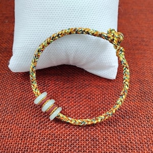 May include: Braided bracelet with a mix of yellow, green, and brown threads. The bracelet features three white, circular beads. The background includes a white pillow and a red textured surface.