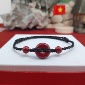 May include: A handmade bracelet featuring a black woven cord with a central, circular, deep red bead and two smaller red beads. The bracelet is displayed on a white surface with a red base, with a blurred background.