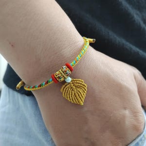 May include: A gold-colored woven bracelet with a leaf charm. The bracelet features a multi-colored woven band with red, blue, and yellow threads. The leaf charm is a detailed, gold-colored design. The bracelet is worn on a wrist.