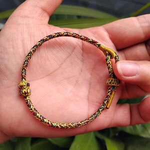 May include: A braided bracelet with a mix of red, yellow, green, and black threads. The bracelet has a knot closure and a loop for adjusting the size. The bracelet is held in a hand.
