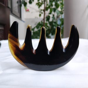 Puede incluir: Un peine de dientes anchos con forma curva de media luna. El peine presenta una superficie lisa y pulida con un patrón jaspeado de vetas de color negro profundo y tonos ámbar translúcidos en todo el material, terminando en cinco dientes.
