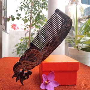 Puede incluir: Un peine de madera marrón oscuro con intrincadas tallas de dragones. El peine presenta dientes finos y un diseño de dragón detallado en el mango. Descansa sobre una caja naranja con una flor morada, sugiriendo un artículo de regalo.