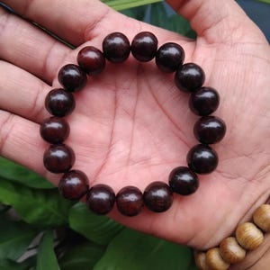 Puede incluir: Una pulsera de cuentas marrón oscuro con cuentas grandes y redondas. La pulsera se sostiene en una mano, mostrando la superficie pulida y lisa de las cuentas. Las cuentas tienen un rico tono marrón rojizo, lo que sugiere un material natural.