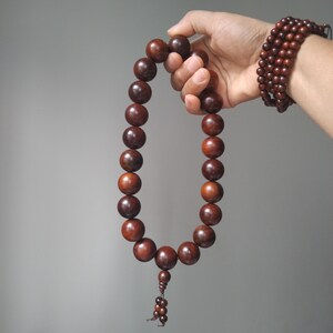 Op de afbeelding: Een streng van grote, gladde, donkerbruine houten kralen in een cirkelvormige lus, met een kleinere kraal en een decoratief kwastje van kleine kraaltjes aan de onderkant. Een bijpassende meerlaagse armband van kleine donkerbruine houten kralen wordt gedragen.