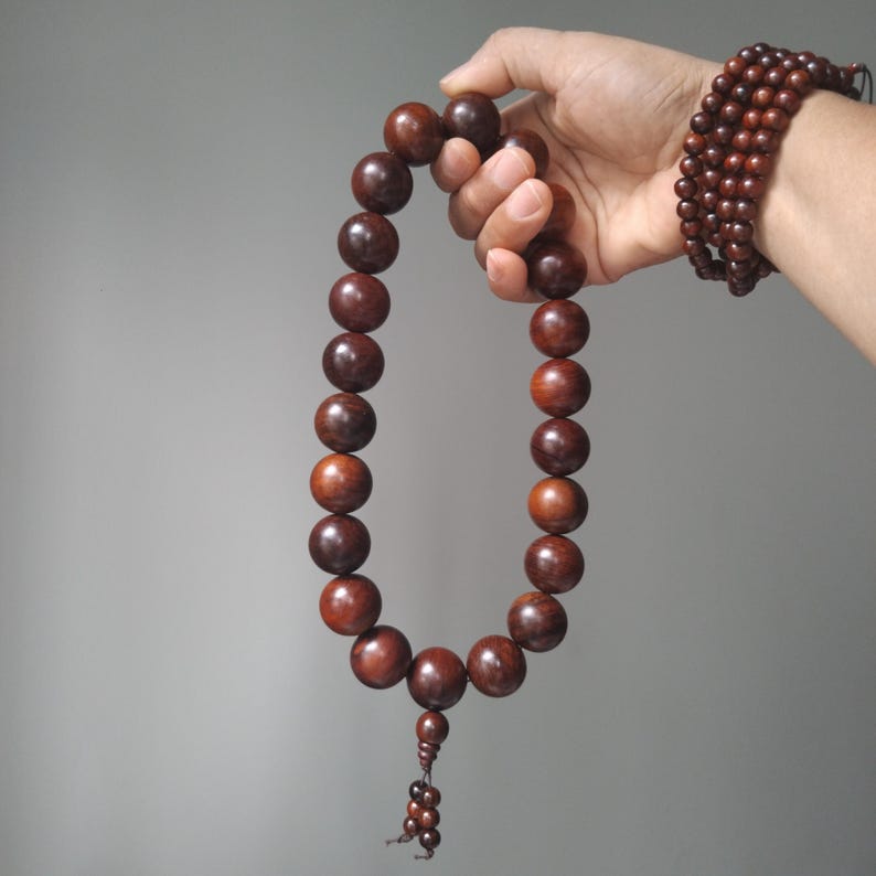 Op de afbeelding: Een streng van grote, gladde, donkerbruine houten kralen in een cirkelvormige lus, met een kleinere kraal en een decoratief kwastje van kleine kraaltjes aan de onderkant. Een bijpassende meerlaagse armband van kleine donkerbruine houten kralen wordt gedragen.