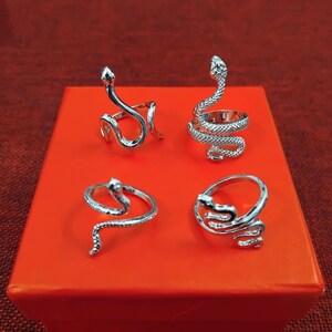 May include: Four silver snake rings on a red box. Each ring has a detailed snake design, with the snake's body wrapping around the finger. The rings vary in style, with some having the snake's head upright. The rings are made of silver.