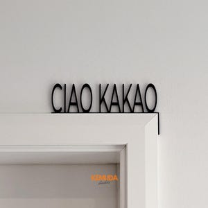 K&ouml;nnte beinhalten: Schwarzes Metallschild mit der Aufschrift "CIAO KAKAO" in einer stilisierten Schriftart, montiert &uuml;ber einem wei&szlig;en T&uuml;rrahmen. Das Schild ist an der Wand und am T&uuml;rrahmen befestigt und dient als dekoratives Element.