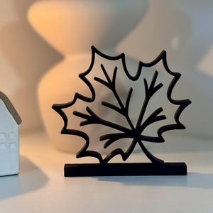 Könnte beinhalten: Schwarze Ahornblatt-Silhouette auf einem kleinen Ständer. Ein weißer Keramik-Kerzenhalter in Hausform mit einer brennenden Kerze und eine weiße Vase mit getrockneten Pflanzen im Hintergrund. Die Objekte stehen auf einer weißen Oberfläche.