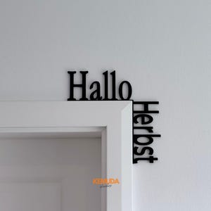 Könnte beinhalten: Ein schwarzes, L-förmiges Wanddekor, das passgenau um die Ecke eines Türrahmens angebracht wird. Das Schild zeigt den deutschen Schriftzug "Hallo" horizontal entlang der Oberkante und "Herbst" vertikal entlang der Seitenkante.