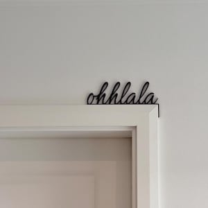 Könnte beinhalten: Schwarze Metall-Wanddekoration mit dem geschwungenen Schriftzug "ohhlala" über einem weißen Türrahmen. Dieses dekorative Element soll die Ästhetik eines Zuhauses oder Büros verbessern.