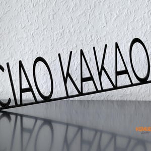 K&ouml;nnte beinhalten: Schwarzes Metallschild mit der Aufschrift "CIAO KAKAO" in einer stilisierten Schriftart. Das Schild ist abgewinkelt und wirft einen Schatten auf die Oberfl&auml;che. Ein dekorativer Artikel.