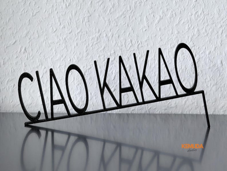 K&ouml;nnte beinhalten: Schwarzes Metallschild mit der Aufschrift "CIAO KAKAO" in einer stilisierten Schriftart. Das Schild ist abgewinkelt und wirft einen Schatten auf die Oberfl&auml;che. Ein dekorativer Artikel.