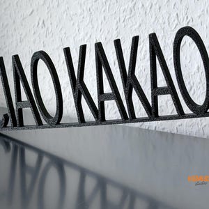 K&ouml;nnte beinhalten: Schwarzes Metallschild mit den Worten "CIAO KAKAO" in einer stilisierten Schriftart. Das Schild steht auf einer dunklen Oberfl&auml;che, mit einer strukturierten wei&szlig;en Wand im Hintergrund. Ein dekorativer Artikel.