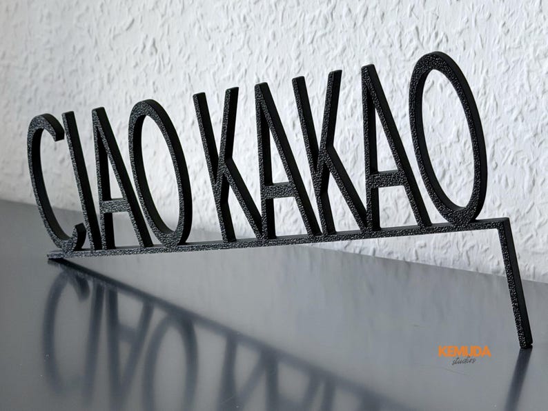 K&ouml;nnte beinhalten: Schwarzes Metallschild mit den Worten "CIAO KAKAO" in einer stilisierten Schriftart. Das Schild steht auf einer dunklen Oberfl&auml;che, mit einer strukturierten wei&szlig;en Wand im Hintergrund. Ein dekorativer Artikel.