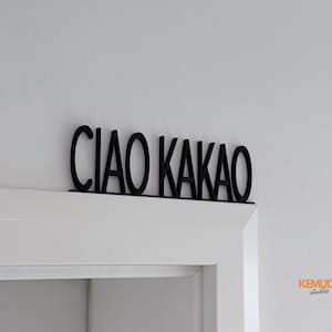 K&ouml;nnte beinhalten: Schwarzes, lasergeschnittenes Schild mit der Aufschrift "CIAO KAKAO" in einer stilisierten Schriftart. Das Schild ist &uuml;ber einem wei&szlig;en T&uuml;rrahmen an einer wei&szlig;en Wand angebracht. Dekorationselement.