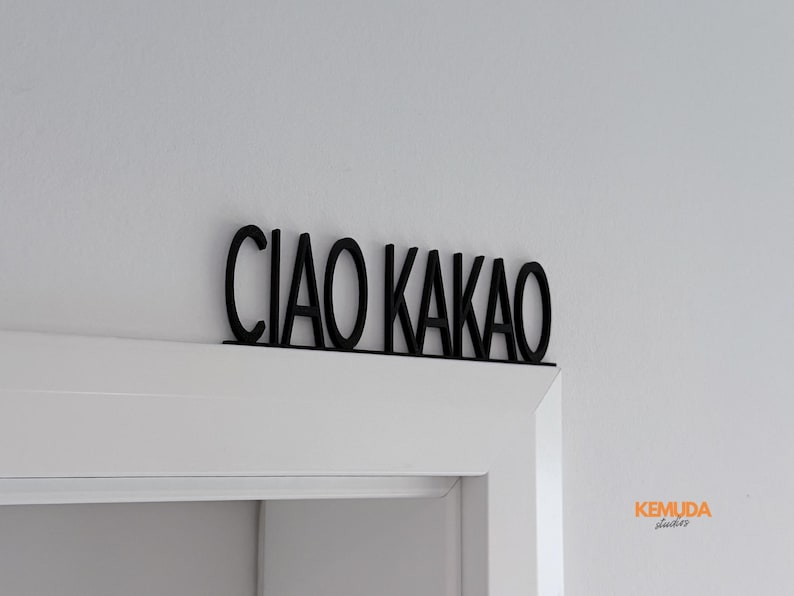 K&ouml;nnte beinhalten: Schwarzes, lasergeschnittenes Schild mit der Aufschrift "CIAO KAKAO" in einer stilisierten Schriftart. Das Schild ist &uuml;ber einem wei&szlig;en T&uuml;rrahmen an einer wei&szlig;en Wand angebracht. Dekorationselement.