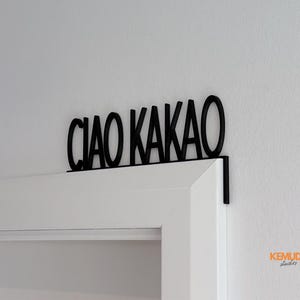 K&ouml;nnte beinhalten: Schwarzes Metallschild mit der Aufschrift "CIAO KAKAO" &uuml;ber einem wei&szlig;en T&uuml;rrahmen. Das Schild, in einer serifenlosen Schriftart, ist mit einer kleinen Halterung am T&uuml;rrahmen befestigt. Der Hintergrund ist eine wei&szlig;e Wand.