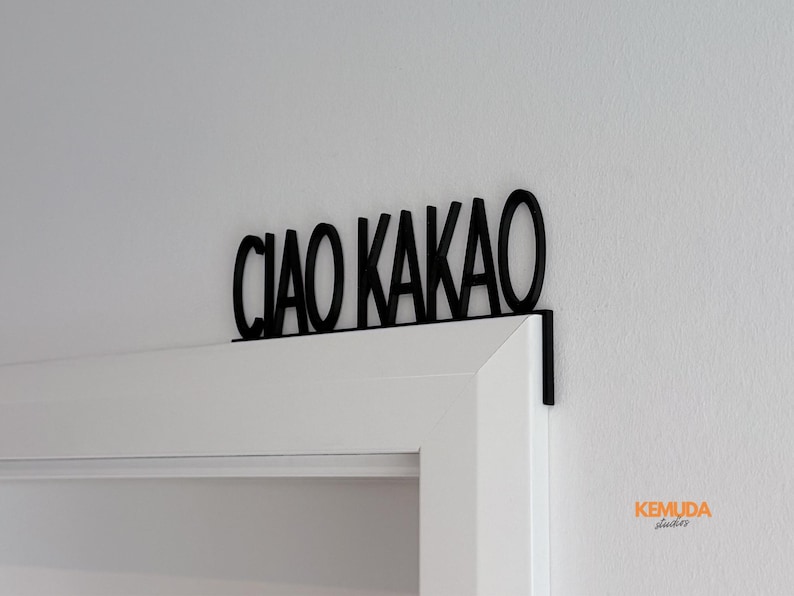 K&ouml;nnte beinhalten: Schwarzes Metallschild mit der Aufschrift "CIAO KAKAO" &uuml;ber einem wei&szlig;en T&uuml;rrahmen. Das Schild, in einer serifenlosen Schriftart, ist mit einer kleinen Halterung am T&uuml;rrahmen befestigt. Der Hintergrund ist eine wei&szlig;e Wand.