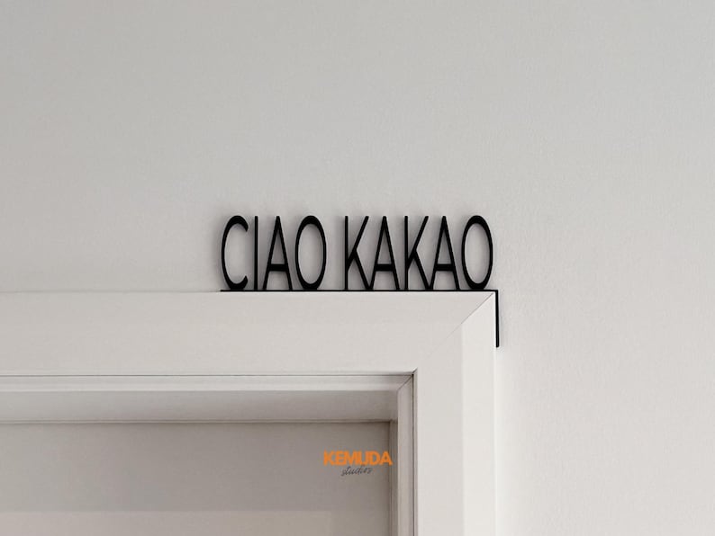 K&ouml;nnte beinhalten: Schwarzes Metallschild mit der Aufschrift "CIAO KAKAO" in einer stilisierten Schriftart, montiert &uuml;ber einem wei&szlig;en T&uuml;rrahmen. Das Schild ist an der Wand und am T&uuml;rrahmen befestigt und dient als dekoratives Element.