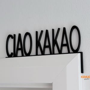 K&ouml;nnte beinhalten: Schwarzes Holzschild mit der Aufschrift "CIAO KAKAO" in fetter, serifenloser Schrift. Das Schild ist &uuml;ber einem wei&szlig;en T&uuml;rrahmen angebracht. Das Schild ist ein dekorativer Artikel.