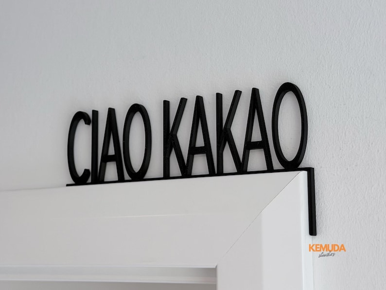 K&ouml;nnte beinhalten: Schwarzes Holzschild mit der Aufschrift "CIAO KAKAO" in fetter, serifenloser Schrift. Das Schild ist &uuml;ber einem wei&szlig;en T&uuml;rrahmen angebracht. Das Schild ist ein dekorativer Artikel.