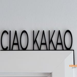 K&ouml;nnte beinhalten: Schwarzes Holzschild mit den Worten "CIAO KAKAO" in einer fetten, serifenlosen Schrift. Das Schild ist &uuml;ber einem wei&szlig;en T&uuml;rrahmen montiert, mit einem kleinen Logo in der Ecke. Ein dekorativer Artikel.