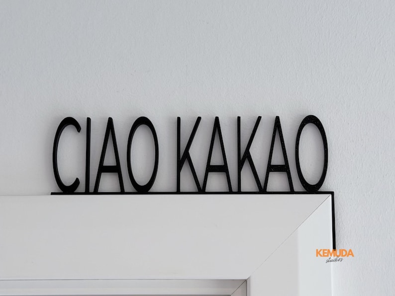 K&ouml;nnte beinhalten: Schwarzes Holzschild mit den Worten "CIAO KAKAO" in einer fetten, serifenlosen Schrift. Das Schild ist &uuml;ber einem wei&szlig;en T&uuml;rrahmen montiert, mit einem kleinen Logo in der Ecke. Ein dekorativer Artikel.