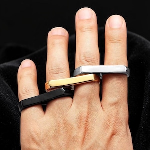 May include: A hand wearing four square-shaped rings in black, gold, and silver. The rings have a flat top and a geometric design. The background is black.