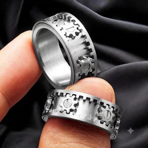 Könnte beinhalten: Ein silberfarbener Metallring mit einem rotierenden Außenband im mechanischen Zahnraddesign. Die Oberfläche des Rings zeigt mehrere kleine zahnradförmige Elemente, die jeweils mit einer horizontalen Linie in der Mitte versehen sind.