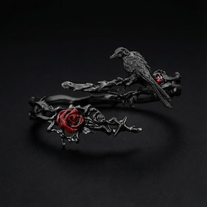 May include: A black metal bracelet featuring a raven perched on a branch with a red rose and red accents. The bracelet has a gothic or fantasy theme, with intricate details and a dark aesthetic.