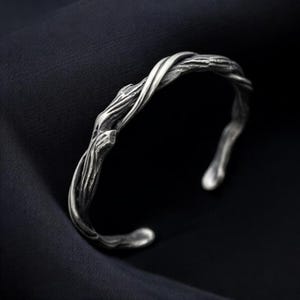 May include: A silver cuff bracelet featuring a textured, organic design that mimics the appearance of intertwined tree branches or gnarled wood. The metal has a darkened, oxidized finish in the recessed areas, highlighting the detailed bark-like texture.