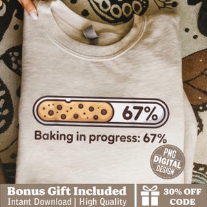 May include: A light beige sweatshirt with a cookie graphic and the text "Baking in progress: 67%". The graphic shows a cookie with chocolate chips and a progress bar. A "PNG DIGITAL DESIGN" sticker is also visible.
