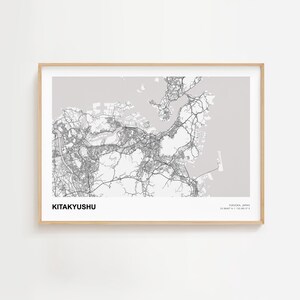 Puede incluir: Una impresión enmarcada del mapa de Kitakyushu, Japón, en blanco y negro. El mapa de la ciudad es detallado, mostrando carreteras y áreas urbanas sobre un fondo gris claro. El texto "KITAKYUSHU" está en la parte inferior izquierda.