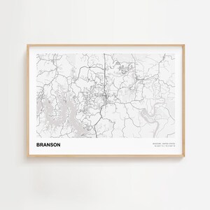 May include: A framed map print of Branson, Missouri, in grayscale. The map details the city's roads, waterways, and surrounding areas. The word "BRANSON" is printed in bold, black letters at the bottom left. The frame is a light wood color.