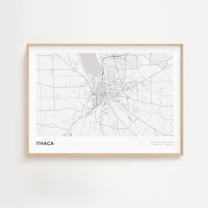 May include: A framed black and white map print of Ithaca, New York, with the city name in a bold font at the bottom left. The map shows roads, buildings, and a lake. The frame is a light wood color.