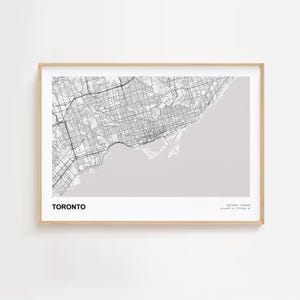 May include: A framed print showing a detailed black and white map of Toronto, Canada. The city's layout is set against a light grey background. The word "TORONTO" is printed in bold black letters at the bottom, with the coordinates below.