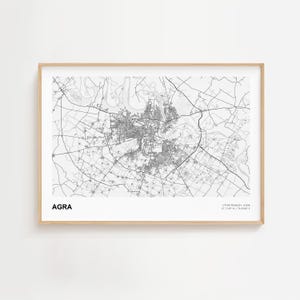 May include: A framed black and white map print of Agra, India. The city map is detailed, showing roads and buildings. The word "AGRA" is printed in a sans-serif font at the bottom left. Coordinates are printed at the bottom right.
