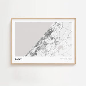 Può includere: Una stampa incorniciata in bianco e nero della mappa di Rabat, Marocco. La mappa mostra la disposizione della città, comprese le strade e le caratteristiche costiere. La parola "RABAT" è stampata in basso a sinistra. La cornice è di colore legno chiaro.