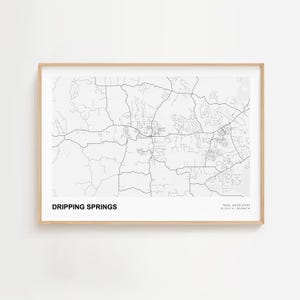 May include: A framed black and white map print of Dripping Springs, Texas, with a detailed road network. The text "DRIPPING SPRINGS" is at the bottom, with the state and coordinates below. The frame is a light wood color.