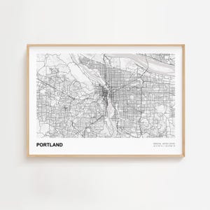 Może przedstawiać: Oprawiona czarno-biała mapa Portland w stanie Oregon, z nazwą miasta wydrukowaną pogrubioną czcionką na dole. Mapa jest szczegółowa, pokazując ulice i drogi wodne. Rama jest w jasnym kolorze drewna.