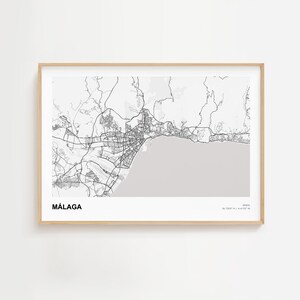May include: A framed black and white map of Málaga, Spain. The map shows the city's layout, including roads, buildings, and the coastline. The word "MÁLAGA" is printed in bold, black letters at the bottom left. The frame is a light wood color.
