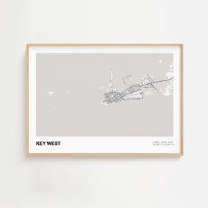May include: A framed print featuring a detailed map of Key West, Florida, in black and white. The map is set against a light gray background, with the words "KEY WEST" at the bottom left. The frame is a light wood color.
