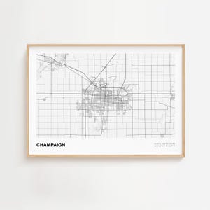 May include: A framed black and white map of Champaign, Illinois, United States. The map shows the city's layout with roads and buildings. The word "CHAMPAIGN" is printed in black at the bottom left. The frame is a light wood color.