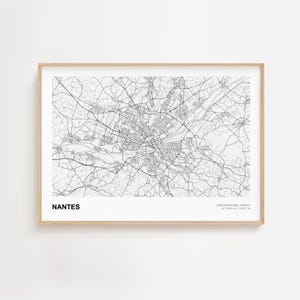 May include: A framed black and white map of Nantes, France. The map details the city's road network and waterways. The city name is printed in a bold, sans-serif font at the bottom left. The frame is a light wood color.