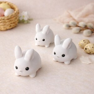 May include: Three white ceramic bunny figurines with black eyes and small smiles are arranged on a light-coloured surface. The bunnies have rounded bodies and upright ears. Easter-themed decor.