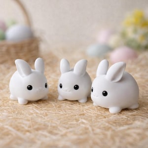 Peut inclure: Trois figurines de lapins blancs avec des yeux noirs et de petits nez sont disposées sur un lit de paille. Les lapins ont des corps arrondis et des oreilles droites, ce qui leur donne une apparence mignonne. L'arrière-plan comprend un panier et des œufs aux couleurs pastel.