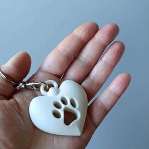 Puede incluir: Un llavero blanco en forma de coraz&oacute;n con un dise&ntilde;o de huella de pata recortado en el centro. El coraz&oacute;n est&aacute; unido a un mosquet&oacute;n de metal plateado y a una anilla circular, lo que permite sujetarlo de forma segura a llaves o bolsos.