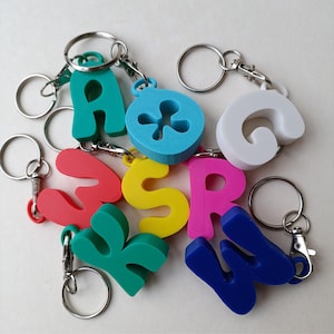 Könnte beinhalten: Eine Sammlung bunter Schlüsselanhänger in Buchstabenform. Die Schlüsselanhänger sind in verschiedenen Farben erhältlich, darunter Grün, Rot, Gelb, Pink, Blau und Weiß. Jeder Buchstabe ist an einem silbernen Schlüsselring mit einem Verschluss befestigt.