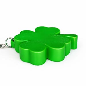 May include: A bright green, four-leaf clover keychain. The clover is made of a solid, glossy material and has a rounded shape. A silver-colored chain is attached to the clover. The background is white.