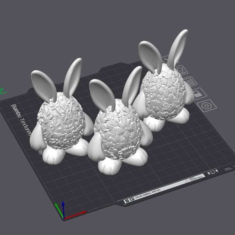 May include: Three white bunny figurines with detailed floral patterns on their bodies and long ears. The bunnies are standing on a dark gray surface, showcasing a 3D-printed design. The overall aesthetic is clean and modern.